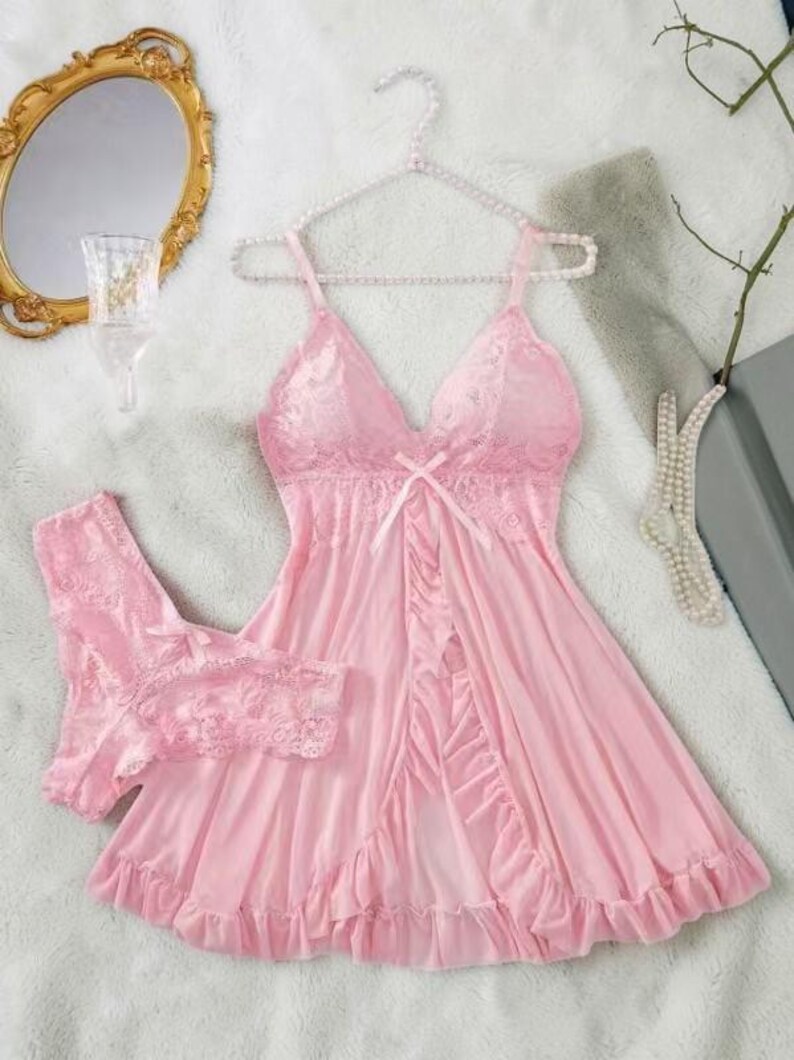 A vibrant pink lace lingerie set laid flat against a soft, textured background features a sheer nightdress with a deep v-neckline, delicate floral lace cups, thin shoulder straps, and a front bow accent alongside matching lace panties.