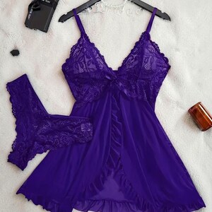 A vibrant purple lace lingerie set laid flat against a soft, textured background features a sheer nightdress with a deep v-neckline, delicate floral lace cups, thin shoulder straps, and a front bow accent alongside matching lace panties.