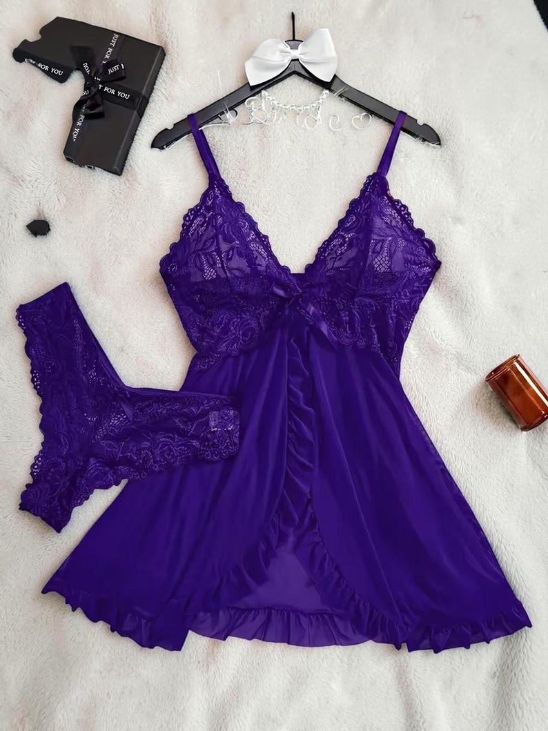 A vibrant purple lace lingerie set laid flat against a soft, textured background features a sheer nightdress with a deep v-neckline, delicate floral lace cups, thin shoulder straps, and a front bow accent alongside matching lace panties.