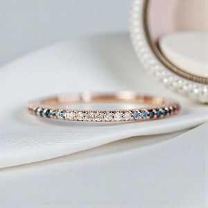 Puede incluir: Anillo de banda de oro rosa con una fila de piedras preciosas blancas y azules alternas. El anillo está sobre una superficie de satén blanco, con un elemento decorativo borroso en el fondo.