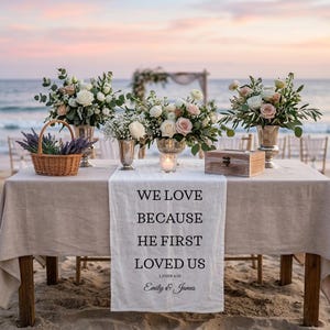 May include: A beach wedding reception table is set with floral arrangements in silver vases, a wooden box, and a woven basket. A white table runner displays the text "WE LOVE BECAUSE HE FIRST LOVED US" and the names "Emily & James".