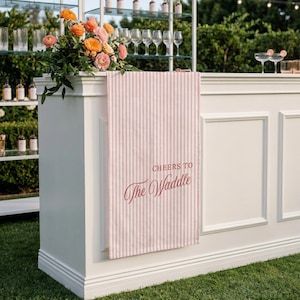 May include: A white bar with a pink and white striped towel that reads "Cheers to The Waddle." The bar is decorated with flowers and glassware. The bar is set up outdoors on a green lawn.