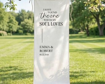 May include: A white banner with the text "I HAVE FOUND the ONE WHOM MY SOUL LOVES" in black script. Below the text are the names "EMMA & ROBERT" and the date "06.22.2026". The banner is hanging from a tree branch.