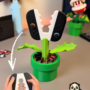 Könnte beinhalten: Ein grüner, pflanzenförmiger Controller-Halter und Aufbewahrungsständer mit weißem, gezacktem Oberteil und Schlitzen für Spielkassetten im Sockel. Der Text ZUBEHÖR PASSEND FÜR NINTENDO SWITCH 2 ist oben im Bild zu sehen.