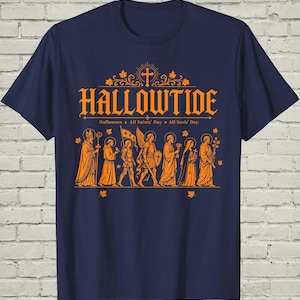 May include: A navy blue short-sleeve t-shirt featuring an orange graphic print. The design displays the text HALLOWTIDE, Halloween, All Saints' Day, All Souls' Day, and a row of seven figures in robes and armor with halos, accented by maple leaves.