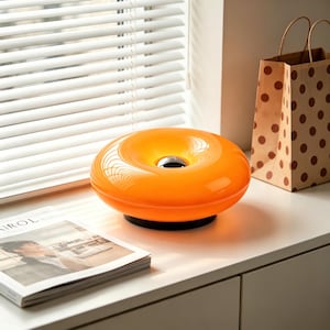 May include: An orange, donut-shaped table lamp with a black base sits on a white surface. A magazine and a brown paper bag with polka dots are also visible. The lamp has a glossy finish and a central, dark-colored element.