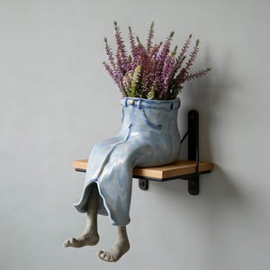 May include: A whimsical ceramic planter shaped like a pair of blue jeans, complete with pockets and a button. The planter is filled with purple flowers and sits on a wooden shelf. The legs of the planter dangle, adding a playful touch.