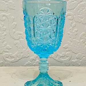 May include: A vintage, light blue glass goblet with a detailed, textured pattern. The goblet features a starburst design in the center and a textured rim. The stem and base are also intricately designed, creating a classic, elegant look.