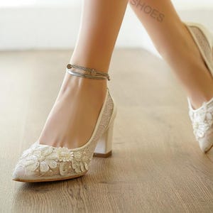 May include: Ivory lace heels with floral embellishments and a block heel. The pointed-toe shoes feature a delicate lace overlay and a silver ankle bracelet. The text "SMYRNALPSHOES" is visible on the leg. These elegant shoes are suitable for special occasions.