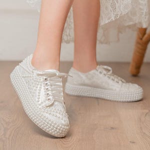 Könnte beinhalten: Weiße Sneaker mit Perlenbesatz. Die Schnürschuhe sind mit kleinen, runden Perlen bedeckt, mit Perlendetails auf den Sohlen. Die Sneaker sind für Bräute oder besondere Anlässe konzipiert. Modische Schuhe.
