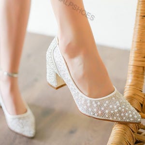 May include: White, embellished block heel pumps with a pointed toe. The shoes are covered in small, sparkling details, with a mesh overlay. The heels are approximately 2 inches high. The shoes are part of a bridal or formal wear collection.