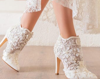 May include: White lace ankle boots with high heels. The boots feature intricate floral lace detailing and are adorned with small pearls. The heels are a light beige colour, and the boots are set against a light background.