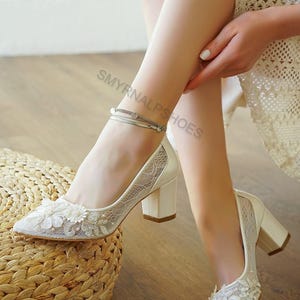 May include: Ivory-colored bridal heels with a floral lace overlay and a block heel. The pointed-toe pumps feature delicate floral embellishments and a silver ankle bracelet. The shoes are on a wooden floor.
