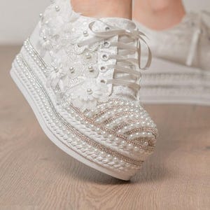 May include: White lace platform sneakers with pearl and rhinestone embellishments. The sneakers have a lace-up closure and are decorated with floral appliques. The platform sole is approximately 2 inches high. The text "SMYRNALPSHOES" is visible on the leg.