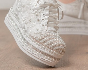 May include: White lace platform sneakers with pearl and rhinestone embellishments. The sneakers have a lace-up closure and are decorated with floral appliques. The platform sole is approximately 2 inches high. The text "SMYRNALPSHOES" is visible on the leg.