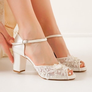 Puede incluir: Zapatos de novia de satén blanco con tacón ancho y correa en el tobillo. El diseño de punta abierta presenta delicados encajes y adornos florales. Los tacones tienen una altura de aproximadamente 7.6 cm.