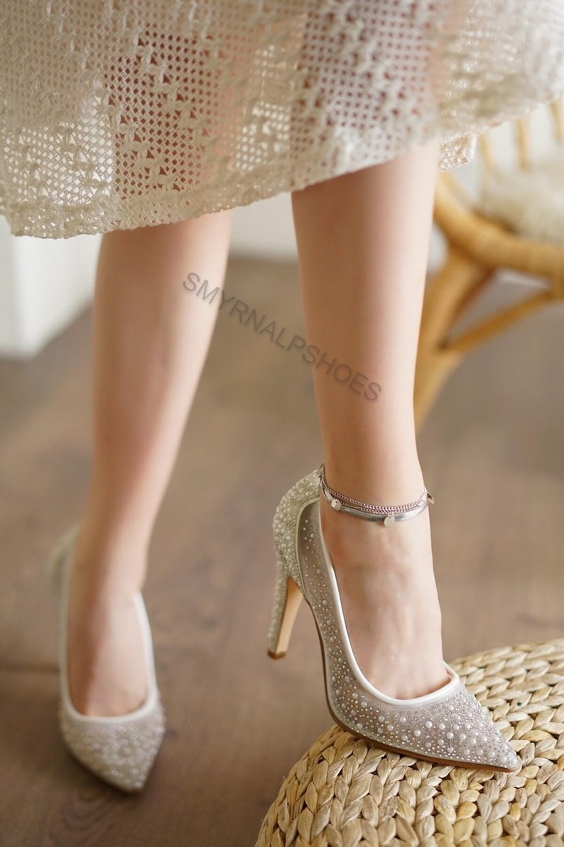 May include: Silver high heels with pointed toes and embellished with rhinestones. The heels have a delicate ankle strap and are set against a neutral background. The text "SMYRNALPSHOES" is visible.
