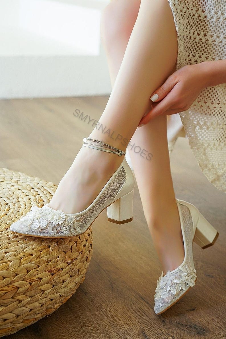 May include: Ivory-colored bridal heels with a floral lace overlay and a block heel. The pointed-toe pumps feature delicate floral embellishments and a silver ankle bracelet. The shoes are on a wooden floor.