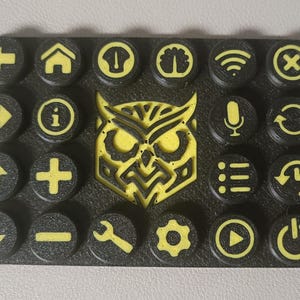 May include: Black rectangular control panel with yellow icons and an owl design. The panel features various symbols like arrows, a house, a plus sign, and a power button. The owl is in the center.