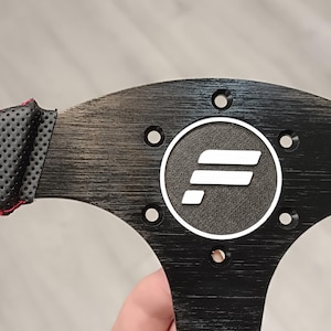 Puede incluir: Un volante negro con un agarre de cuero perforado y costuras rojas. El centro presenta un emblema circular con un logotipo blanco. Los radios del volante tienen un aspecto de metal cepillado.