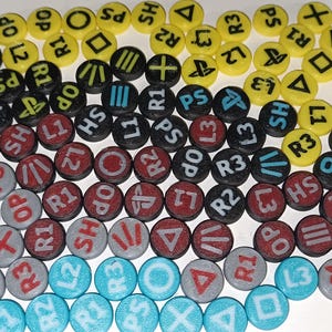 Può includere: Una collezione di bottoni rotondi e colorati con vari simboli e testo, tra cui forme e lettere come "R1", "L3" e "PS". I bottoni sono gialli, neri, rossi, grigi e blu.