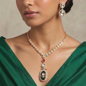 Puede incluir: Un conjunto de collar y pendientes colgantes en tono dorado. El collar presenta una cadena de piedras circulares que conduce a un colgante central con una gran piedra ovalada de color verde oscuro, una pequeña piedra roja y cristales claros.