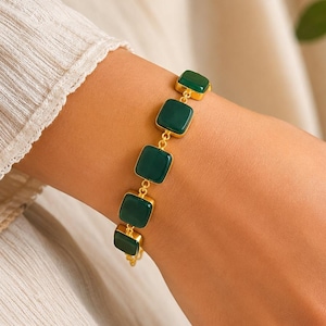 Peut inclure: Un bracelet à chaîne doré composé de cinq pierres carrées vert foncé à surface lisse, serties dans des montures métalliques dorées individuelles. Le bracelet est assemblé par de petits anneaux circulaires dorés reliant chaque monture carrée.