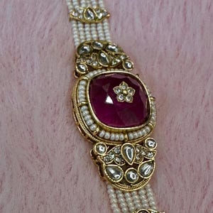 May include: A gold necklace featuring a large, square, ruby-coloured gemstone. The gemstone is framed by small pearls and gold filigree. Multiple strands of small, white pearls form the necklace's base, creating a luxurious look.