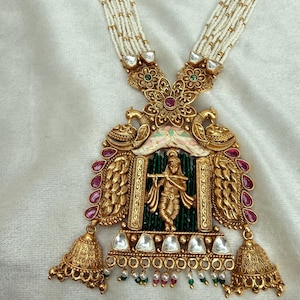 Può includere: Una collana color oro con un grande ciondolo centrale che raffigura una figura che suona il flauto, incorniciata da motivi a pavone, pietre rosse e accenti bianchi. Il ciondolo è affiancato da due ciondoli a forma di campana e fili di perline.