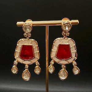 May include: Gold-coloured earrings featuring a red central stone, framed by a white border and clear, teardrop-shaped dangles. The earrings are displayed on a gold stand against a dark background.