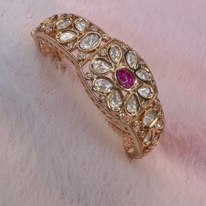 May include: A gold-coloured bracelet with a floral design. The bracelet features clear, teardrop-shaped stones and a central, oval-shaped, ruby-coloured stone. The bracelet is set on a pink background.