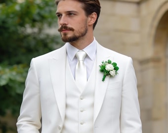 May include: A crisp white three-piece suit consisting of a tailored blazer, a matching button-down vest, and trousers. The ensemble includes a white dress shirt, a solid white necktie, and a small floral boutonniere with white roses and green leaves.