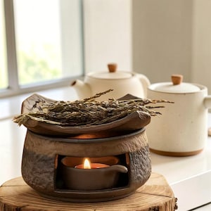 May include: A ceramic tea warmer with a lit candle, topped with dried tea leaves. The warmer sits on a wooden base, with a teapot and a mug in the background. The warmer is brown and has a rustic design.