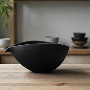May include: A black ceramic bowl with an unusual, flowing shape. The bowl has a curved edge and a small spout on one side. It is placed on a light wooden surface, with other bowls in the background. The bowl is likely for serving or display, and is made of ceramic.