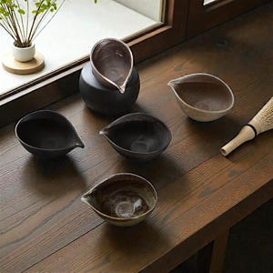 May include: A selection of handcrafted ceramic tea bowls in shades of brown and black, each with a spout. The bowls are arranged on a wooden surface, with a bamboo whisk, suggesting a tea ceremony. The bowls are made of ceramic.