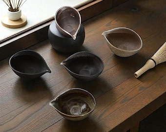 May include: A collection of handcrafted ceramic tea bowls in various shades of brown and black, each with a distinctive spout. The bowls are arranged on a wooden surface, alongside a bamboo whisk, suggesting a tea ceremony setting.