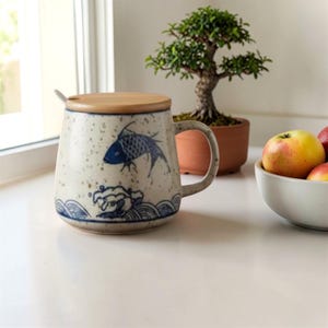 Puede incluir: Una taza de cerámica con tapa de madera y una cuchara pequeña. La taza presenta un diseño de pez azul y olas sobre un fondo blanco moteado. Un bonsái y un cuenco de manzanas están en el fondo.