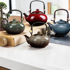 Może przedstawiać: Cztery ceramiczne czajniki w odcieniach zieleni, czerwieni, niebieskiego i brązu. Każdy czajnik ma zaokrąglony korpus, zakrzywioną wylewkę i uchwyt z tkanego materiału. Czajniki są wystawione na białej powierzchni.