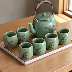 May include: A celadon green tea set comprising a teapot with a woven handle and six matching cups. Each piece is decorated with a delicate floral design in red and brown. The set is presented on a rectangular bamboo tray.