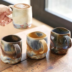 May include: Handmade ceramic mugs in a variety of colours and textures. One mug, held by a hand, contains a latte with a heart design. The other mugs are on a wooden surface, displaying unique shapes and earthy tones.