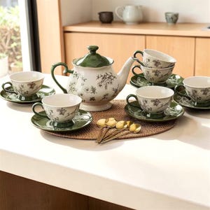 May include: A white and green floral tea set, including a teapot, teacups, and saucers. The set features a green lid and handles, with gold spoons on a woven mat. The teacups and saucers are approximately 10 cm in diameter.