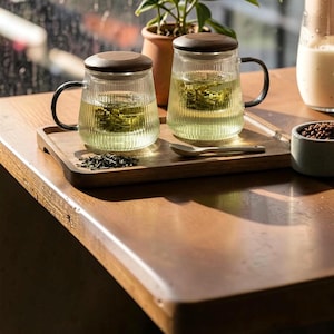May include: Two glass tea mugs with wooden lids and black handles sit on a wooden tray. The mugs are filled with tea and tea leaves. A small ceramic bowl with brown granules and a spoon are also on the tray.
