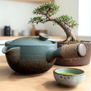 May include: A teal ceramic teapot with a side handle wrapped in twine, a matching lid, and a small bowl of green tea. A bonsai tree in a brown pot sits in the background, creating a serene, natural aesthetic.