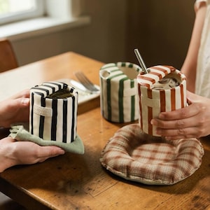 Puede incluir: Tres recipientes de cerámica a rayas en blanco y negro, verde y blanco, y rojo y blanco. Un recipiente tiene una pajita de metal. Los recipientes están sobre una mesa de madera con un paño verde y un posavasos a cuadros.