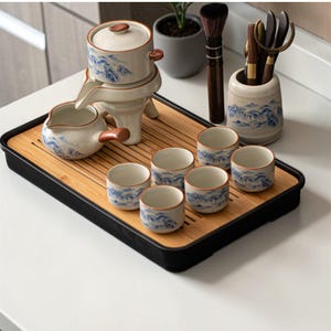 May include: A traditional tea set on a bamboo tray with a black edge. The set includes a teapot, a server, six small cups, and a container for tea utensils. The porcelain items are white with blue mountain designs and brown detailing.