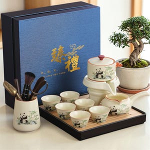May include: A panda-themed tea set, including a teapot, cups, and a serving tray. The set is presented in a blue gift box with gold Chinese characters. A small bonsai tree is in the background. The tea set is designed for serving tea.