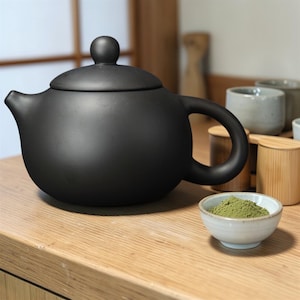 May include: A matte black ceramic teapot with a rounded body, a curved handle, and a lid with a small knob. A small white bowl contains green powder, likely matcha, and is next to the teapot. Several small teacups are in the background.