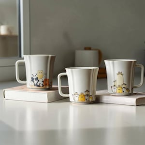 Puede incluir: Tres tazas de cerámica blanca con un diseño ondulado, cada una con un grupo diferente de animales de dibujos animados. Las tazas están colocadas encima de un libro de color claro. Las tazas tienen un asa y son de diferentes tamaños.