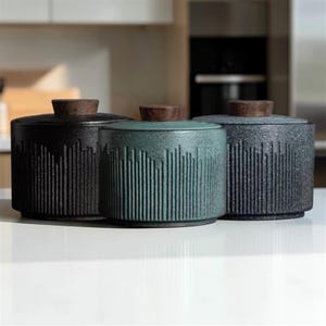 Puede incluir: Tres botes de cerámica con tapas de madera. Los botes son negros, turquesa y grises, cada uno con un diseño de líneas verticales texturizadas. Las tapas son de color madera oscura, sobre una superficie blanca.
