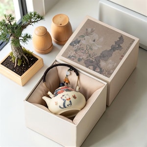 May include: A cream-colored ceramic teapot with a cat design, presented in a beige gift box with floral artwork. The teapot has a dark handle and is accompanied by two wooden containers and a small bonsai tree in a wooden planter.